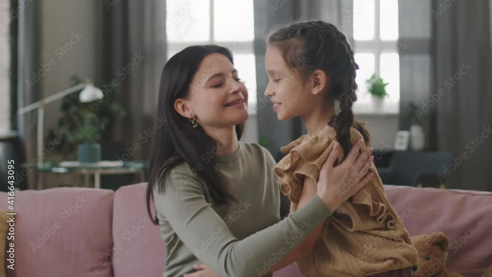 Slowmo panning medium close up of happy attractive woman sitting on sofa in living room hugging 8-year-old daughter smiling to each other gently Slowmo panning medium close up of happy attractive woman sitting on sofa in living room hugging 8-year-old daughter smiling to each other gently