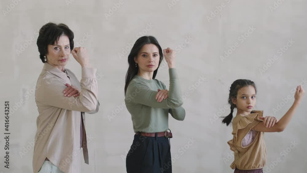 Slow-motion medium studio portrait of three multigenerational women standing in superhero pose at white background, placing one hand on arm, looking at camera seriously Slow-motion medium studio portrait of three multigenerational women standing in superhero pose at white background, placing one hand on arm, looking at camera seriously