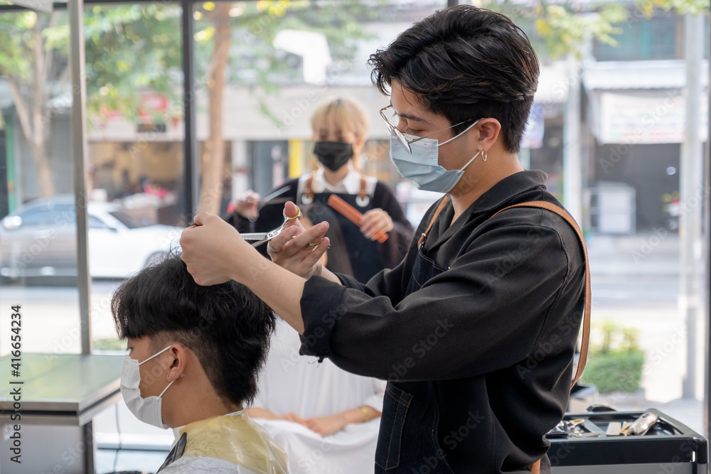 Beauty salon Asian barber man with hygiene mask use scissors cut hair of customer in the shop with other co-worker also take care hair style of woman. Beauty bussiness for good appearance concept. Beauty salon Asian barber man with hygiene mask use scissors cut hair of customer in the shop with other co-worker also take care hair style of woman. Beauty bussiness for good appearance concept.