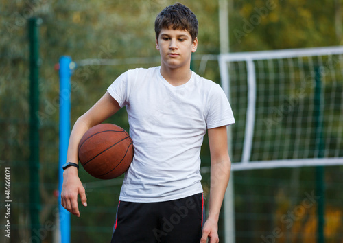Cute young boy plays basketball on street playground in summer. Teenager in white t-shirt with orange basketball ball outside. Hobby, active lifestyle, sports activity for kids.