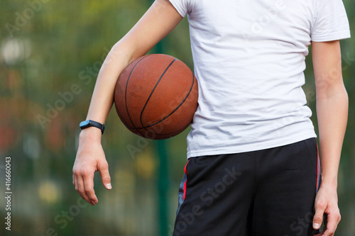 Cute Teenager in white t-shirt with orange basketball ball plays basketball on street playground in summer. Hobby, active lifestyle, sports activity for kids.	
