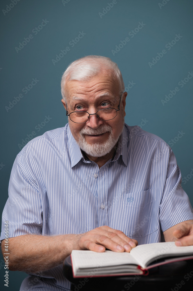 handsome elderly man in glasses holds an open book and looks inquiringly behind the camera, emotions in the frame, gray-blue background, studio shooting, portrait handsome elderly man in glasses holds an open book and looks inquiringly behind the camera, emotions in the frame, gray-blue background, studio shooting, portrait