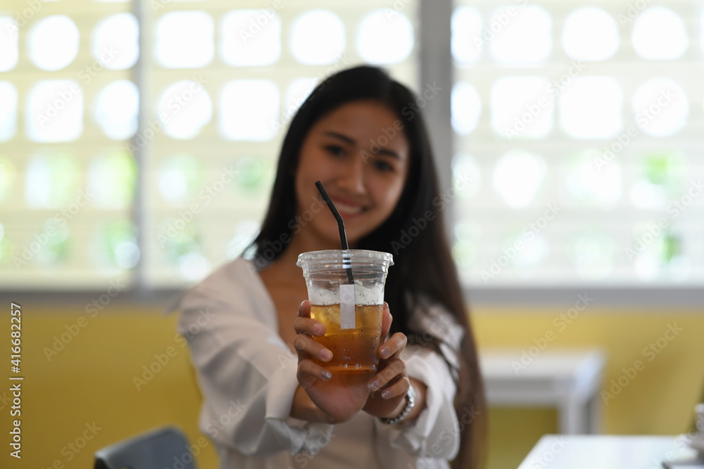 Cheerful young woman holding and showing plastic glass of iced coffee and smiling to camera.