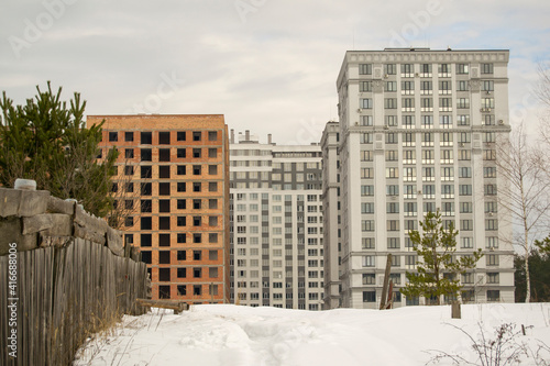 two houses standing side by side. one of the houses is unfinished. snow covered earth. wooden fence leads to houses © Maksym Om