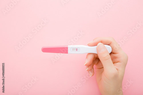 Young adult woman hand holding pregnancy test with one stripe on light pink table background. Pastel color. Negative result. Closeup. Top down view.
