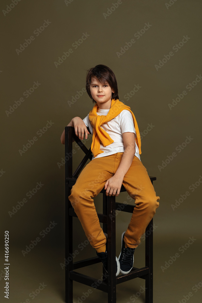 Handsome fashion young boy sits on a chair in studio on green background. Casual style. Stylish shooting in studio Handsome fashion young boy sits on a chair in studio on green background. Casual style. Stylish shooting in studio