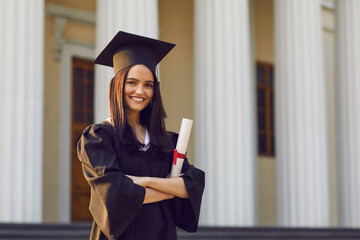 Portrait of confident female student with diploma in hands on university background. Girl is dressed in mortar boards and bachelor mantle. Education, graduation and people concept.