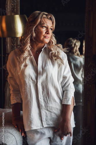 Portrait photo cute middle aged 45 years old blonde woman in domestic room. Female in white clothes in dark interior background. Barefoot in front of mirror. Indoors positive confident mature woman