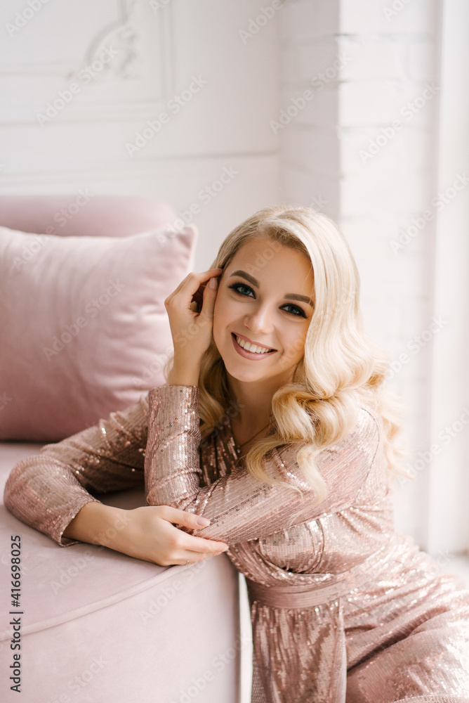Naklejka premium Attractive young blonde woman with curly hair and makeup in a sequined dress in the bright interior of the studio house