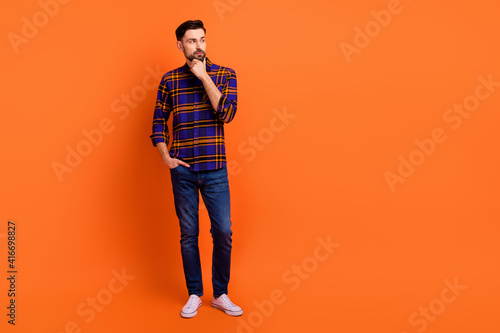 Full length photo of handsome guy arm on chin look interested empty space isolated on orange color background