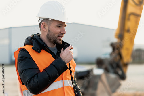Young engineer talking radio communication (walkie talkie) and wearing a white helmet and construction orange vest. Close up engineers working on a building site with the sunny background.