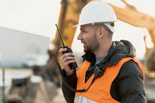 Young engineer talking radio communication (walkie talkie) and wearing a white helmet and construction orange vest. Close up engineers working on a building site with the sunny background.