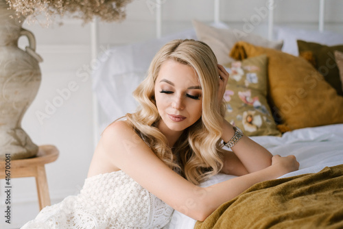 Attractive young blonde woman with curly hair and makeup in a white dress in the bright interior of the studio house