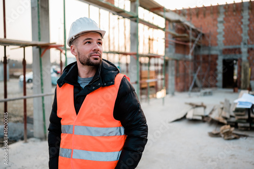 Civil young engineer or architect with white hardhat and orange construction vest on construction site checking progress.