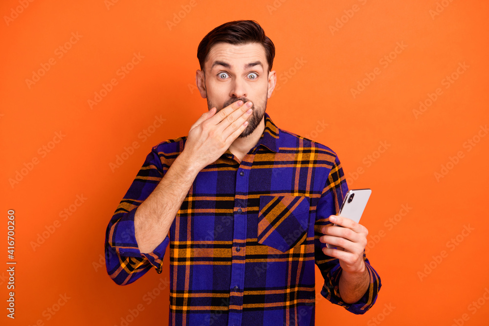 Portrait of handsome guy hold phone arm palm covering mouth lips look camera isolated on orange color background Portrait of handsome guy hold phone arm palm covering mouth lips look camera isolated on orange color background