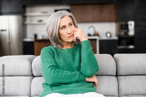 Thoughtful aged woman with grey hair, thinking about the future, planning life, worried about her children, home and property, touching the face, looking aside, sitting on the pure sofa at apartment