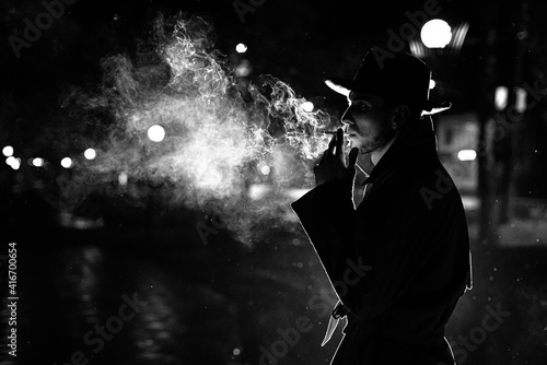 Canvas Print dark silhouette of a man in a hat Smoking a cigarette in the rain on a night str