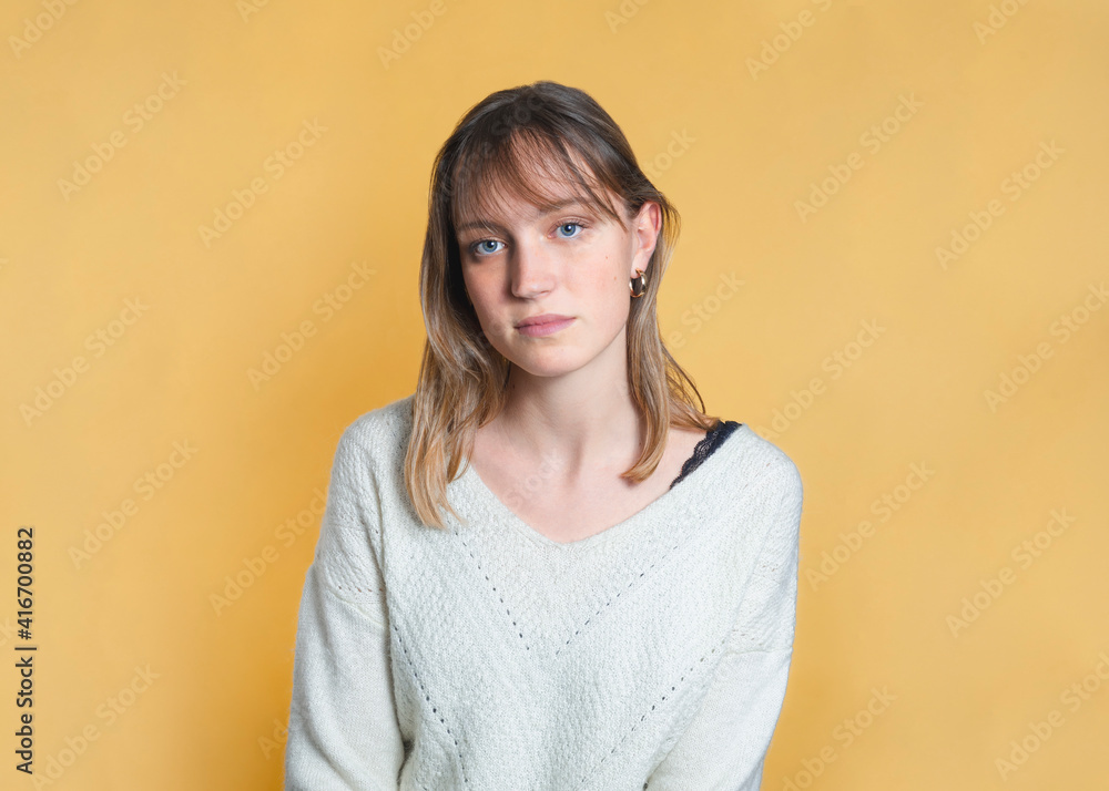 Studio portrait of a woman. She has blond hair, a fringe and blue eyes. She looks serious. She is standing in the center of the image, in front of a yellow background. She is wearing earrings. Studio portrait of a woman. She has blond hair, a fringe and blue eyes. She looks serious. She is standing in the center of the image, in front of a yellow background. She is wearing earrings.