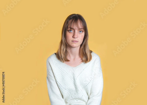 Studio portrait of a woman. She has blond hair, a fringe and blue eyes. She looks serious. She is standing in the center of the image, in front of a yellow background. © Lucille MD