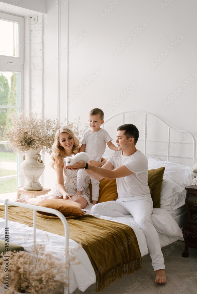 Beautiful young family man woman and son in white clothes play on the bed with a rabbit at home Beautiful young family man woman and son in white clothes play on the bed with a rabbit at home