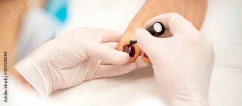Closeup of hands in white gloves applying burgundy nail polish with a brush on woman's toes in a nail salon