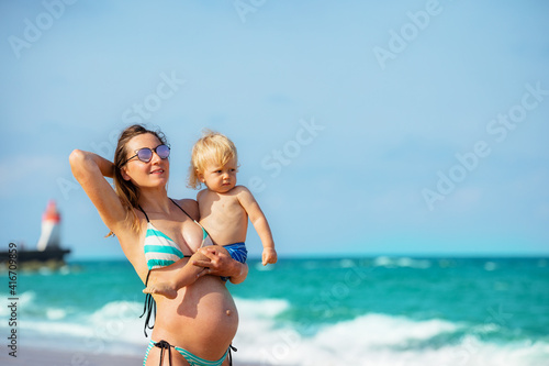 Close portrait of the pregnant mother hold blond toddler standing near sea with lighthouse on background