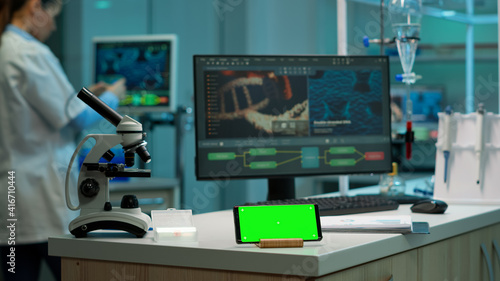 Scientist standing at monitor in background analysing virus evolution while phone with mock-up green screen working in front. Woman lab researcher examining vaccine developent bringing blood samples