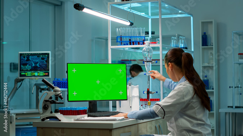 Microbiologist working on computer with display, green chroma key screen talking with coworker about new vacutainer with blood samples. Team of biotechnology scientists developing drugs.