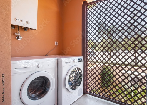 Simple laundry room with a machine for washing and drying, a boiler on top and a fenced off division that shows the outdoor garden. photo