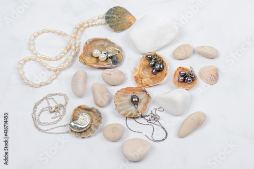 Sea jewelry collection on light marble background with seashells and pebbles. Pearl pendant necklace, black and white pearl earrings and silver chains. Fashion flat lay, ocean summer jewelry