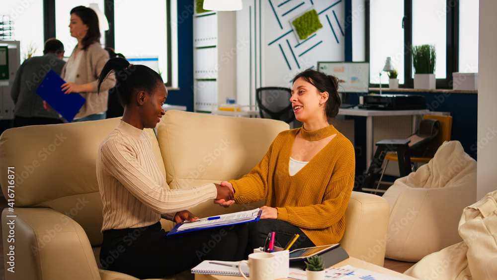 Obraz premium Pleased multiracial business partners sitting on couch in modern office shaking hands after signing partnership contract. Diverse team being happy for successful negotiations in start up company.