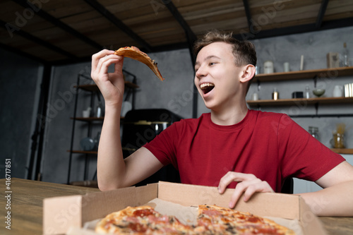 a young guy sitting at a wooden table opened his mouth and is going to send there a piece of pizza he is holding in his hand