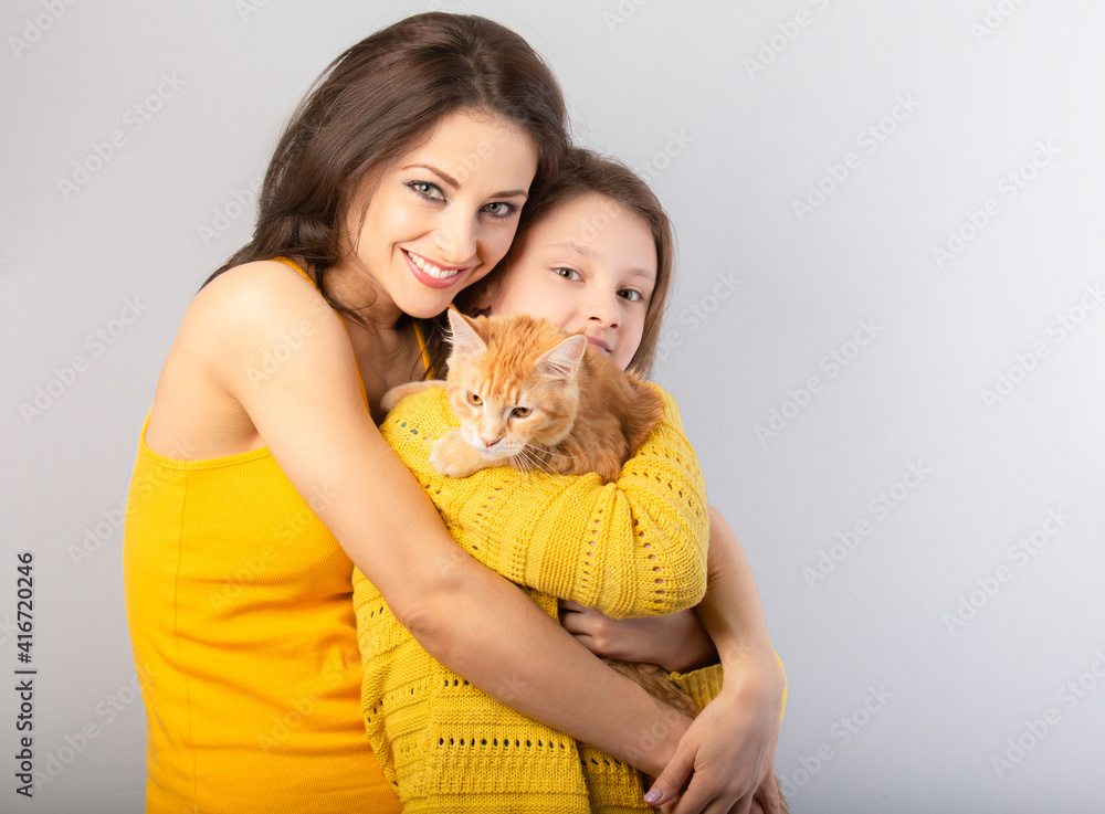Obraz premium Happy young woman hugging her red maine coon kitten and daughter. Closeup portrait of smiling family on blue baclground