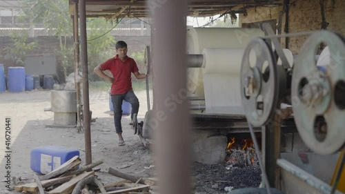 outdoor wide shot of a Indian male local factory labourer standing by running machinery of cloth sheets and looking at camera on a broad daylight photo