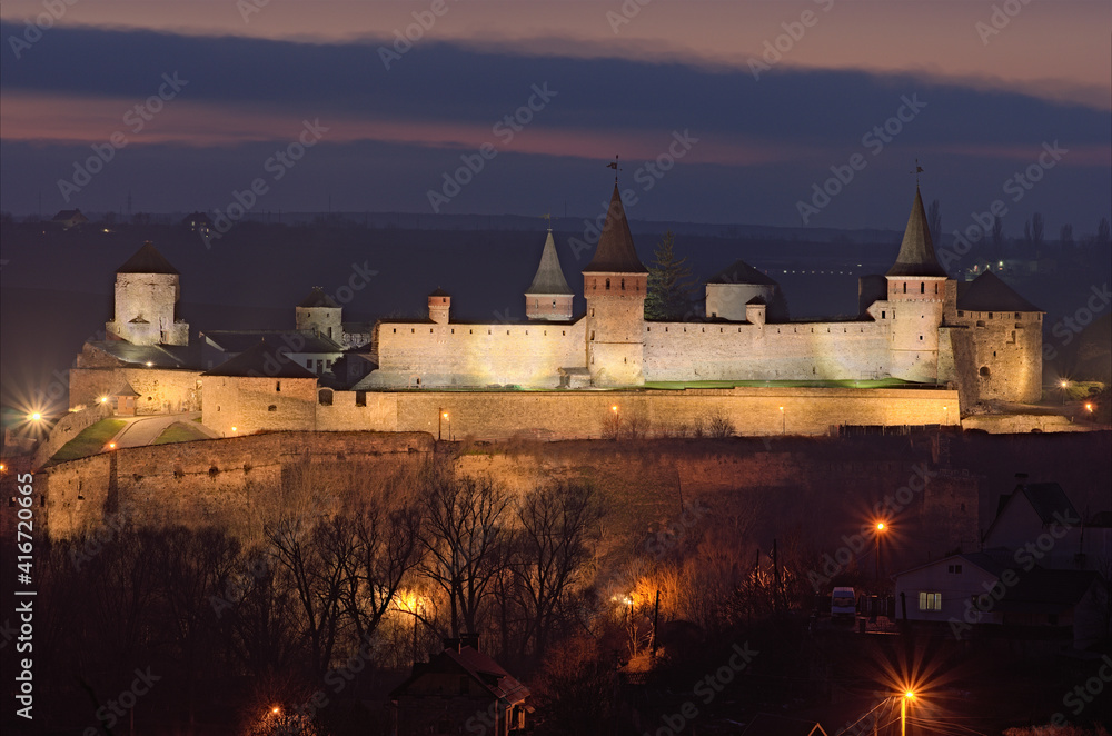 Night light landscape of the medieval Kamianets-Podilskyi castle on the hill near Smotrytsky canyon. Blurred winter sky with clouds in the background. Travel and tourism concept