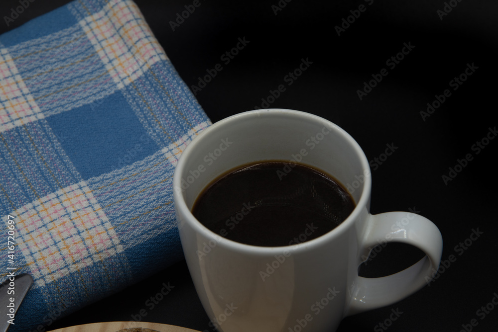 Una taza blanca con cafe, una tostada de pan con un bollo de leche sobre un plato de madera y una mantel azul sobre fondo negro Una taza blanca con cafe, una tostada de pan con un bollo de leche sobre un plato de madera y una mantel azul sobre fondo negro