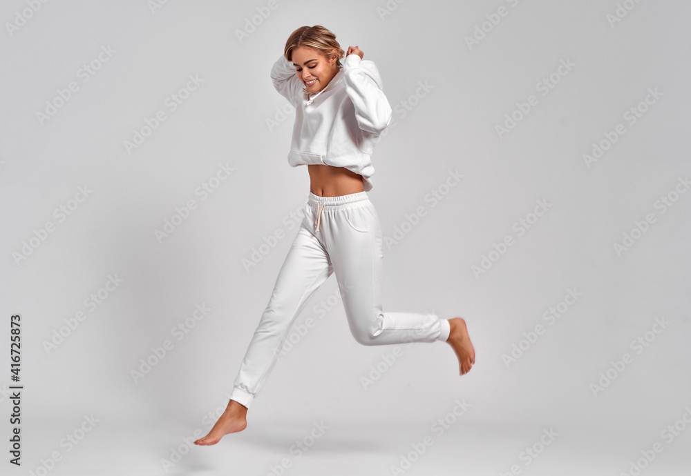 Fototapeta premium Beautiful blonde woman jumping in the air in white tracksuit isolated on gray background