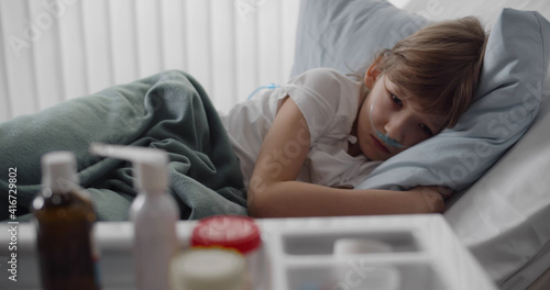 Sick preteen boy with nasal tube lying in hospital bed looking at tray with medication