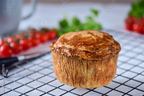 An English pie with minced meat and meat, made from shortcrust pastry, lies on a black metal lattice, side view, in the background cherry tomatoes, greens.