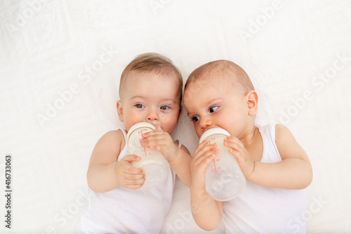 two baby twins boy and girl with a bottle of milk on a white bed at home, baby food concept, place for text © Any Grant