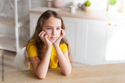 cute school-age girl with curly-haired girls sits at a table in the kitchen, has fun, singing while waiting for food, a delighted child relaxes at the counter, enjoys a happy childhood at home 