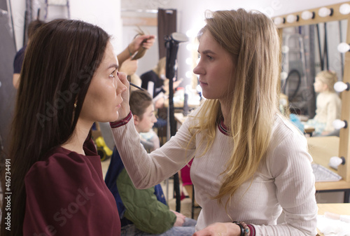 Girl makeup artist, blonde, applying makeup to a girl of 25, European appearance with long dark hair. Process, makeup artist work, beauty industry, generation y. Model preparation in the dressing room