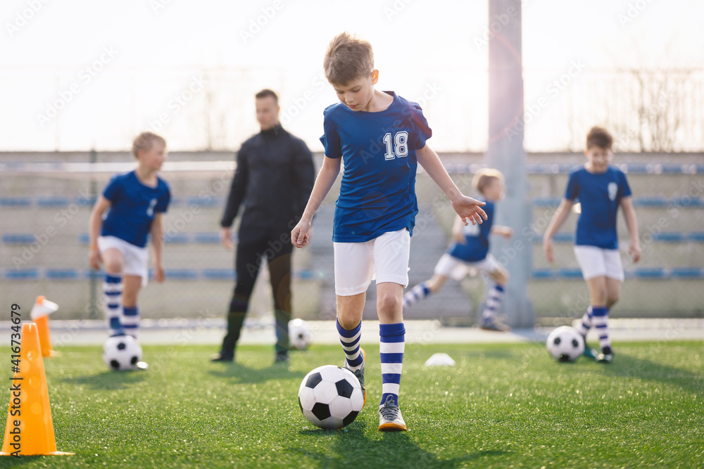 Young Boy Training Sports in Sunny Summer Day. Happy Kids Practicing Soccer on Schoolyard. Young Coach With Children Football Players on Training Session. Sports Stadium Seats in the Background Young Boy Training Sports in Sunny Summer Day. Happy Kids Practicing Soccer on Schoolyard. Young Coach With Children Football Players on Training Session. Sports Stadium Seats in the Background