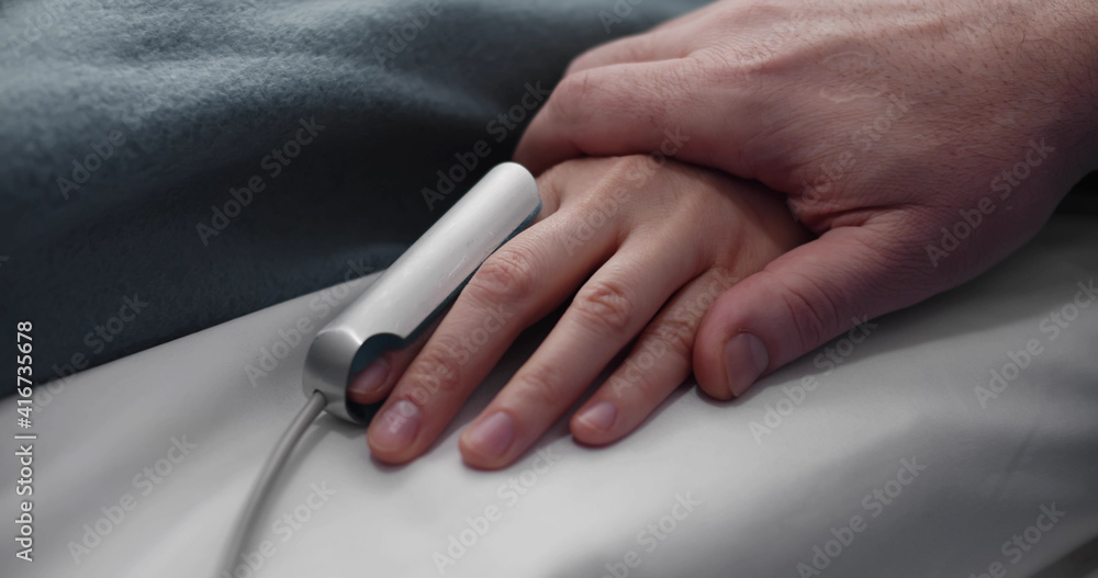 Close up of parent holding hand of sick child with oximeter on finger lying in hospital bed