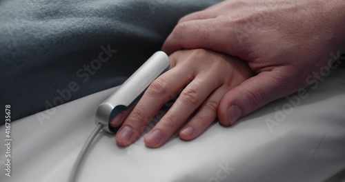 Close up of parent holding hand of sick child with oximeter on finger lying in hospital bed