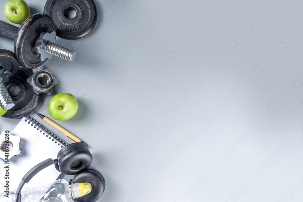 Mens Fitness background with dark Iron dumbbells, sneakers, headphones, gren apples and water bottle. On grey background top view copy space