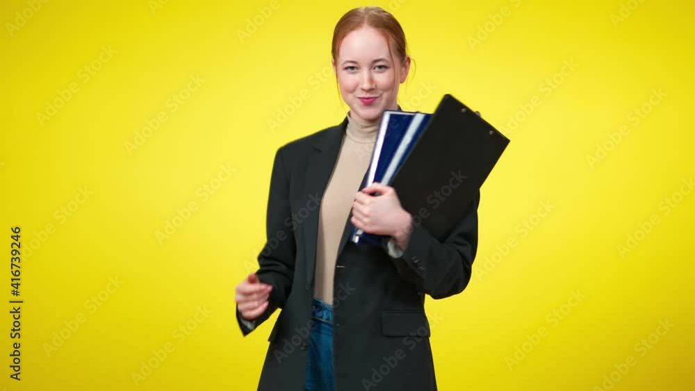 Cheerful confident female CEO with toothy smile dancing with documents at yellow background. Portrait of joyful young slim Caucasian redhead woman rejoicing startup success. Cheerful confident female CEO with toothy smile dancing with documents at yellow background. Portrait of joyful young slim Caucasian redhead woman rejoicing startup success.