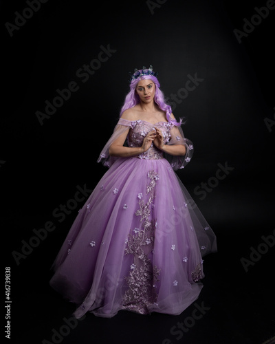 Full length portrait of girl wearing long purple fantasy ball gown with crown and pink hair, standing pose with elegant gestural  movements against a studio background.