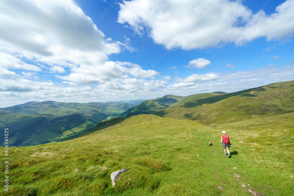 A female hiker descending the summit of  Meall na Aighean towards Glen Lyon with distant views of Ben Lawers on the left in the Scottish Highlands, UK landscapes.