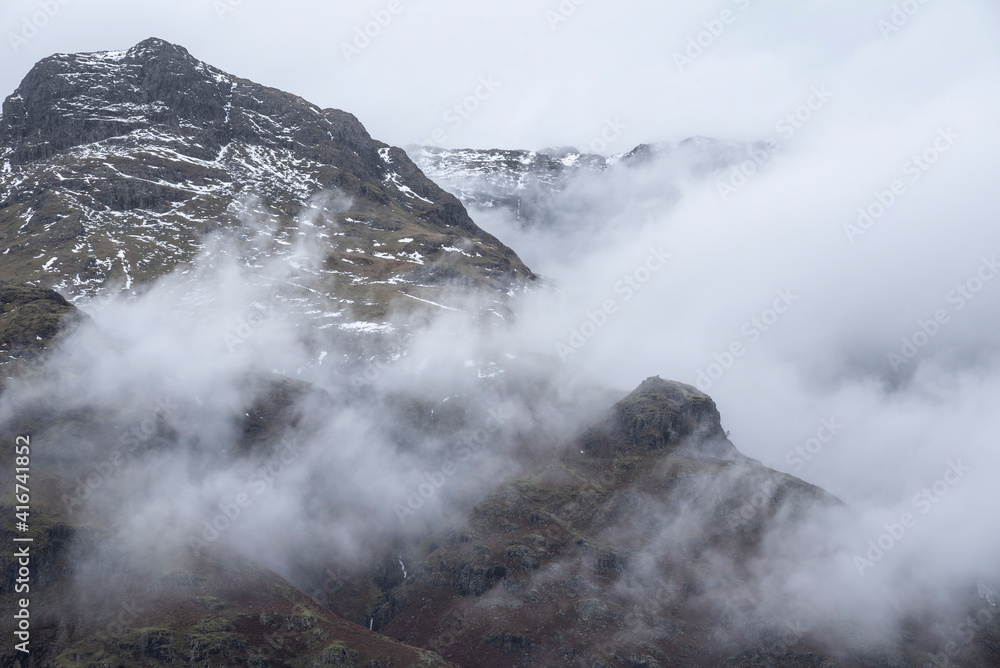Epic Winter landscape image of view from Side Pike towards Langdale pikes with low level clouds on mountain tops and moody mist swirling around